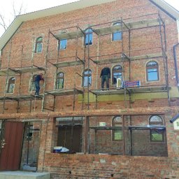 FORTUNA - Dwóch robotników na rusztowaniu przy dwukondygnacyjnym domu z czerwonej cegły z oknami w kształcie łuków, częściowo ocieplonym wełną mineralną.