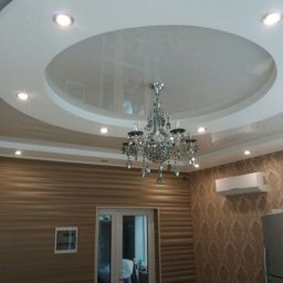 FORTUNA - Nowoczesny salon z podwieszanym, okrągłym sufitem z wbudowanymi punktowymi lampami i kryształowym żyrandolem, ściany wykończone w dwóch różnych wzorach tapet, widoczny klimatyzator i drzwi balkonowe.