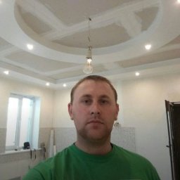 FORTUNA - Mężczyzna w zielonej koszulce na tle sufitu podwieszanego w trakcie montażu, widoczna wisząca żarówka i niedokończone łączenia płyt gipsowo-kartonowych.