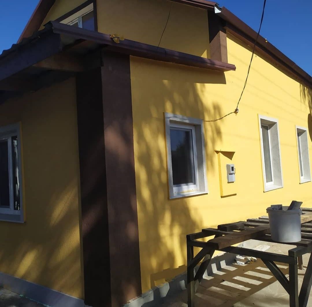 Żółta elewacja domu jednorodzinnego z brązowymi elementami wykończeniowymi, widoczny fragment rusztowania i cień drzewa.