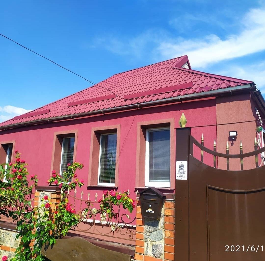 Dom z czerwoną elewacją i dachem z blachodachówki, widoczny z perspektywy ulicy, z dekoracyjnym ogrodzeniem i skrzynką pocztową, przed domem kwitną róże.