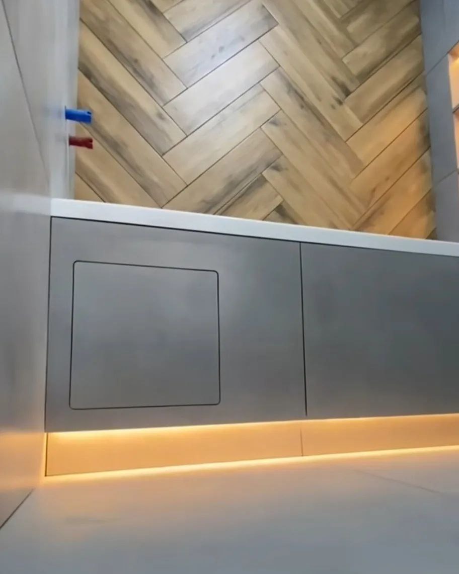 Szara zabudowa pod wanną z ukrytymi drzwiczkami rewizyjnymi, podświetlona listwą LED, na ścianie płytki imitujące drewno ułożone w jodełkę, widoczne rury instalacji wodnej.