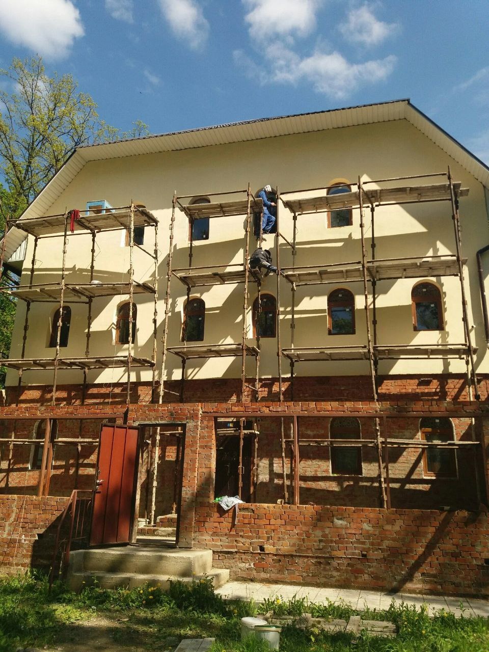 Dwóch pracowników na rusztowaniu podczas prac na elewacji budynku z ceglaną dobudówką, widoczne okna z łukowym wykończeniem i błękitne niebo.