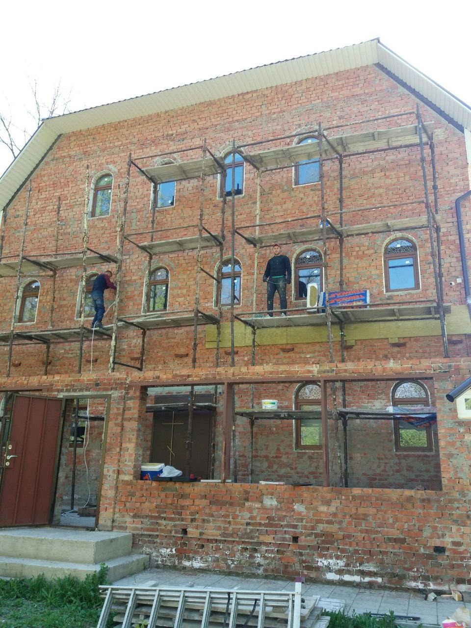 Dwóch robotników na rusztowaniu przy dwukondygnacyjnym domu z czerwonej cegły z oknami w kształcie łuków, częściowo ocieplonym wełną mineralną.