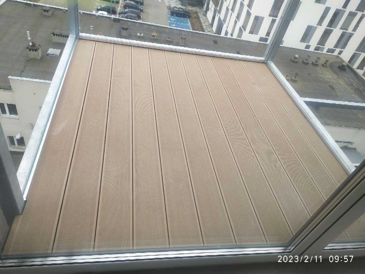 Wykończona podłoga balkonu z desek kompozytowych w odcieniach beżu, widok z góry, panorama miejska w tle, uchwycona przez szybę balkonu.