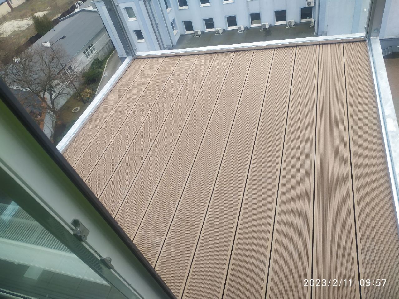 Beżowe deski kompozytowe ułożone na balkonie, widok z góry z perspektywy okna, widoczne elementy mocowania okna, data wykonania zdjęcia w prawym dolnym rogu.
