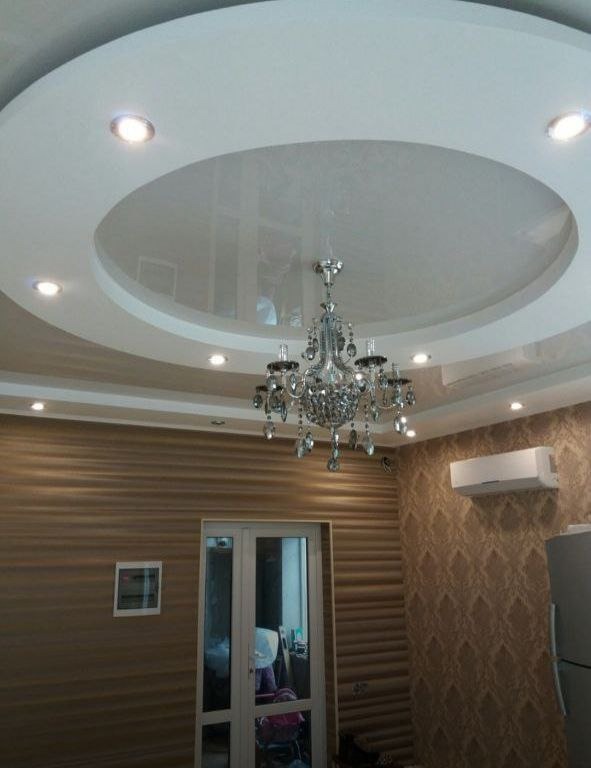 Nowoczesny salon z podwieszanym, okrągłym sufitem z wbudowanymi punktowymi lampami i kryształowym żyrandolem, ściany wykończone w dwóch różnych wzorach tapet, widoczny klimatyzator i drzwi balkonowe.