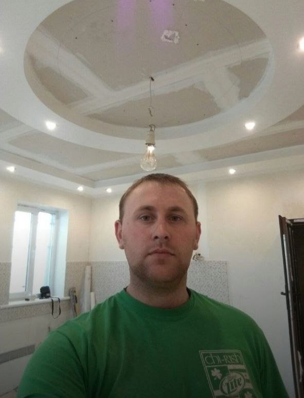 Mężczyzna w zielonej koszulce na tle sufitu podwieszanego w trakcie montażu, widoczna wisząca żarówka i niedokończone łączenia płyt gipsowo-kartonowych.