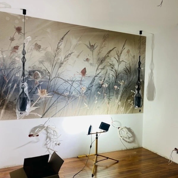 Montaż dwóch czarnych, ozdobnych lamp wiszących na tle ściany z fototapetą przedstawiającą łąkę; jedna lampa jeszcze w folii ochronnej, widoczny statyw oświetleniowy i kable.