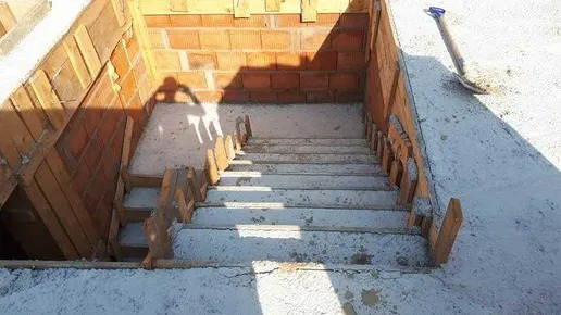 Surowe betonowe schody w trakcie budowy, drewniane szalunki, mur z czerwonej cegły w tle, cień osoby na ścianie, łopata oparta o ścianę po prawej.