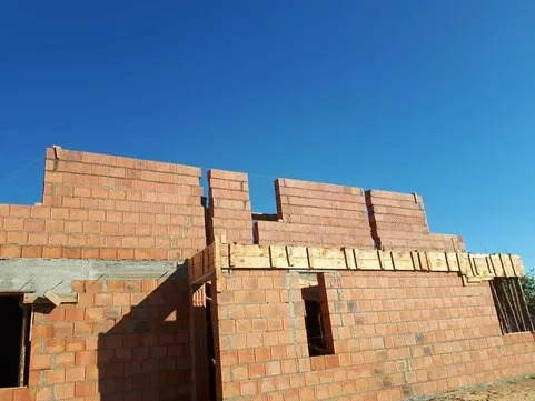 Wznoszenie ścian zewnętrznych z czerwonej cegły na budowie domu jednorodzinnego, widoczne elementy szalunków okiennych i fragmenty zbrojenia, błękitne niebo w tle.