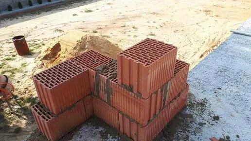 Wznoszenie muru z czerwonych, perforowanych pustaków ceramicznych na fundamencie, widoczna sterta piasku i mieszarka na zewnątrz budynku.