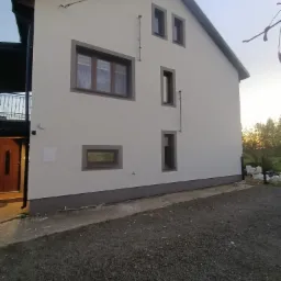 Dwukondygnacyjny dom z jasną elewacją i szarobrązowymi obramowaniami okien oraz ciemną podmurówką, widoczny z boku na żwirowym podłożu.