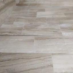 Beżowa, trawertynowa posadzka ułożona w prostokątne płytki, widoczne łączenia i listwa przypodłogowa, ściana wykończona tym samym materiałem.