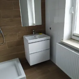 Nowoczesna łazienka z brązowymi płytkami imitującymi drewno, białą umywalką z szafką, grzejnikiem i toaletą w folii ochronnej.