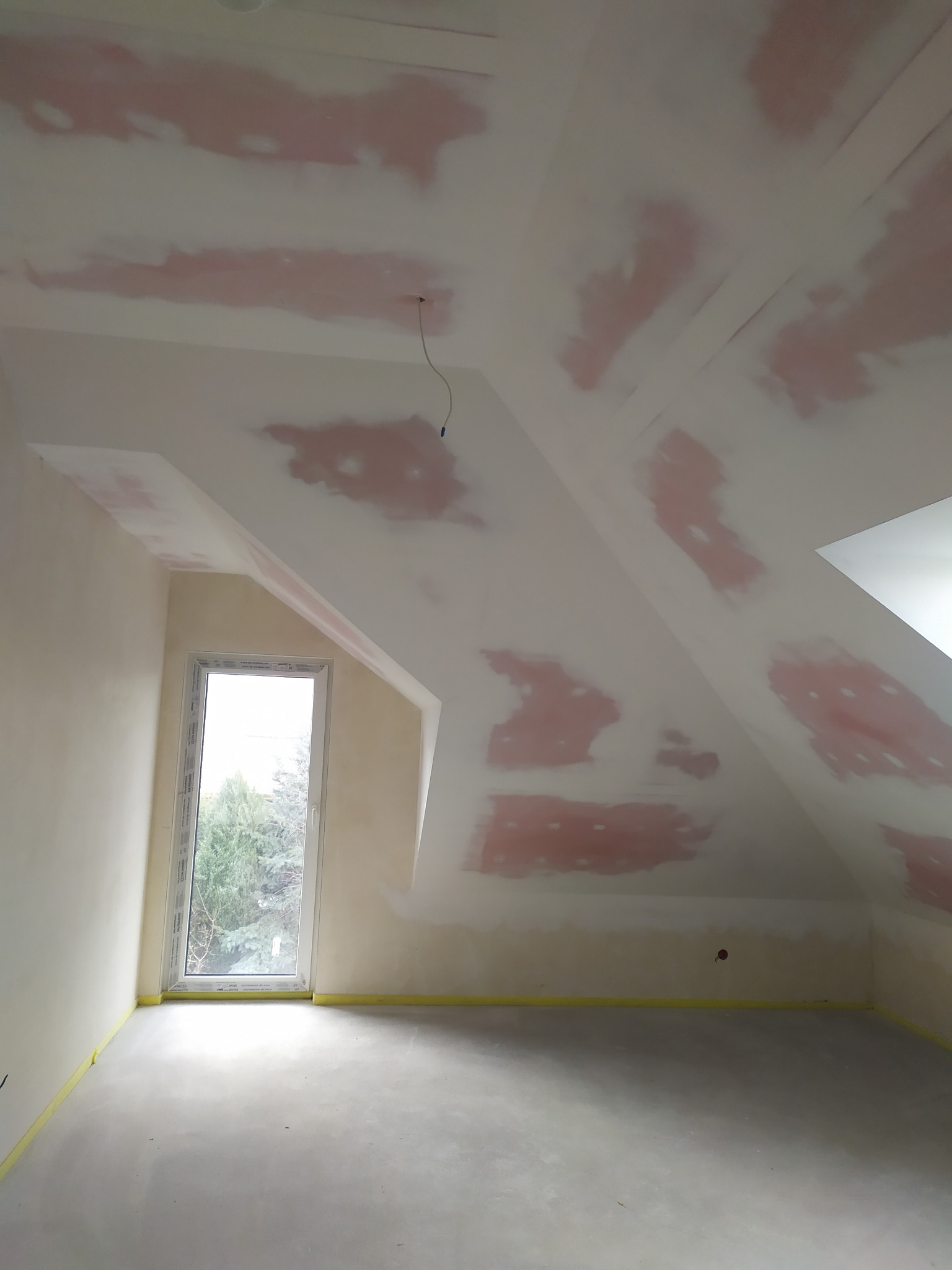 Pokój na poddaszu w trakcie prac przygotowawczych do malowania: zagruntowany sufit ze skosami, oklejone taśmą podłogi, widoczne okno.