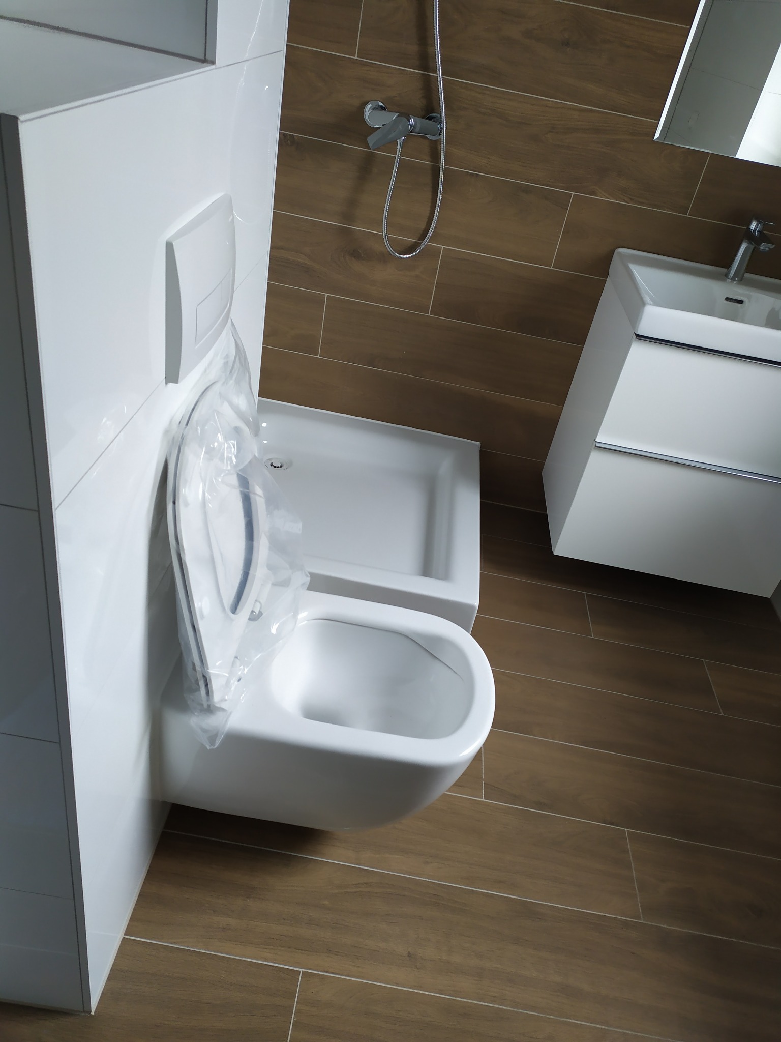Nowoczesna, minimalistyczna łazienka z podwieszaną toaletą jeszcze w folii ochronnej, umywalką nablatową i brązowymi płytkami imitującymi drewno.