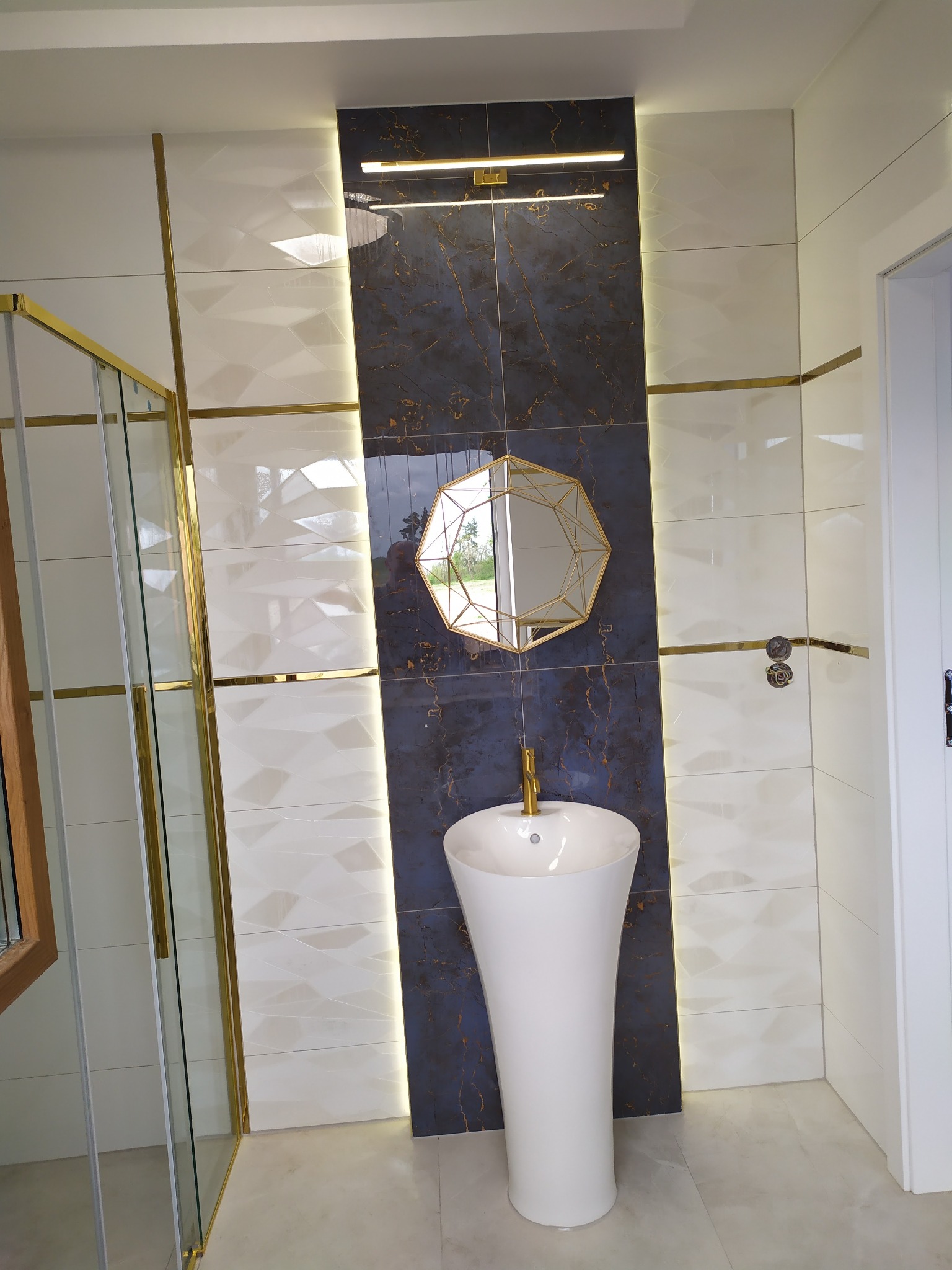 Nowoczesna łazienka z białą umywalką wolnostojącą, lustrem w złotej ramie o kształcie wielokąta i płytkami w odcieniach bieli i granatu z akcentami złota.