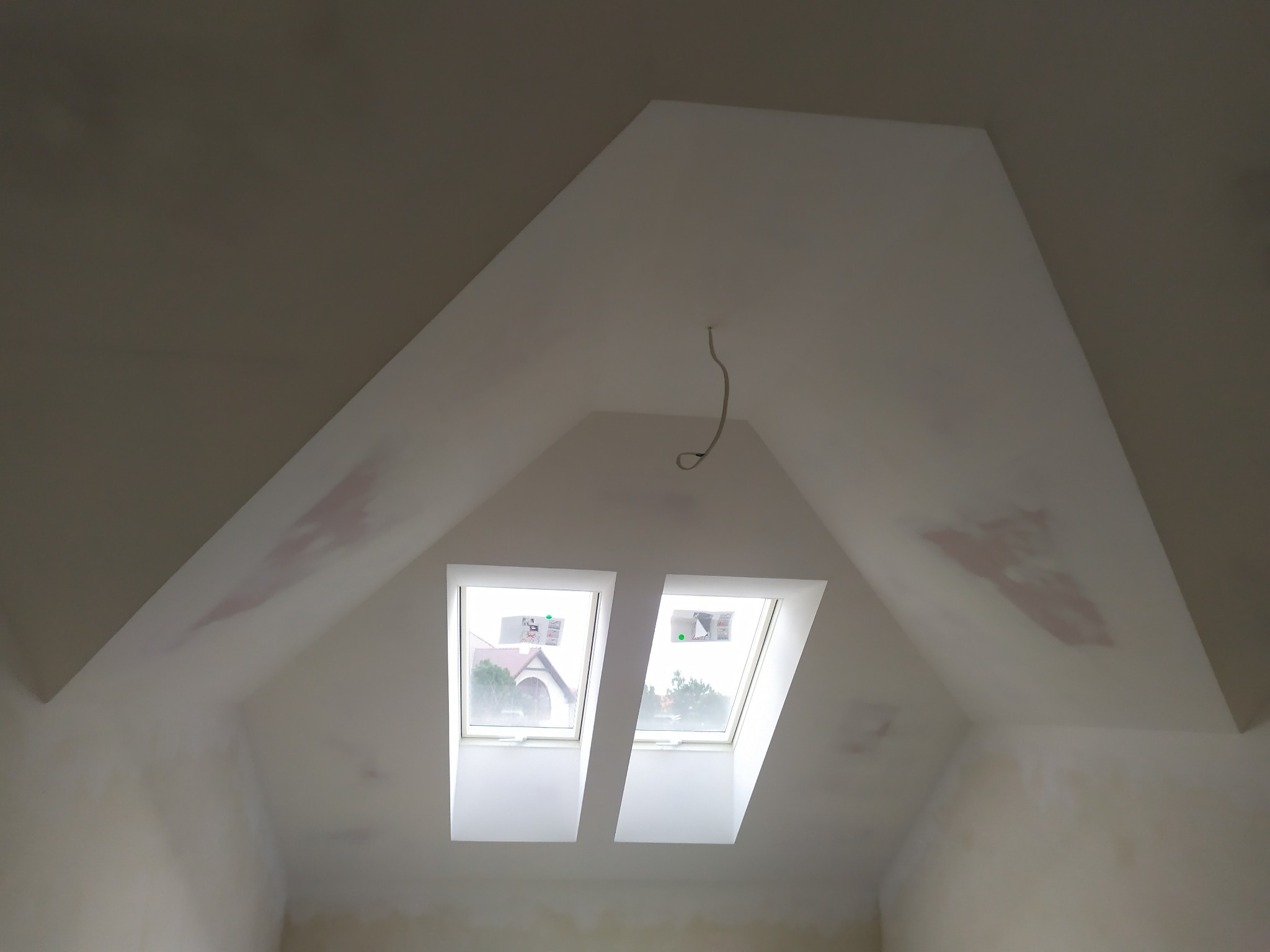 Poddasze w trakcie prac wykończeniowych, widoczne dwa okna dachowe, ślady po łatach i wiszący kabel elektryczny.