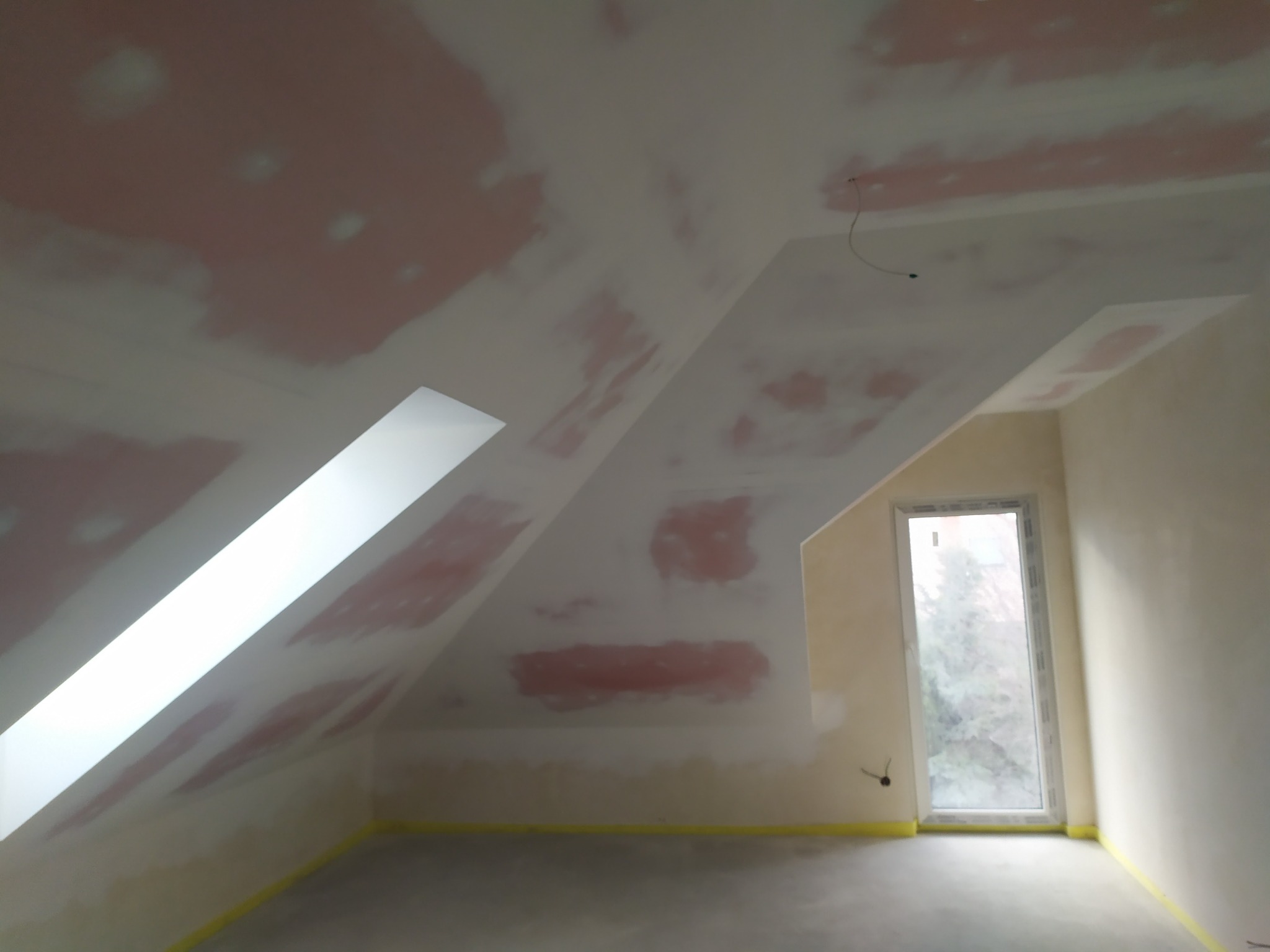 Poddasze w trakcie szpachlowania, widoczne ślady po szpachli na skośnym suficie, zabezpieczona taśmą podłoga i okno dachowe.