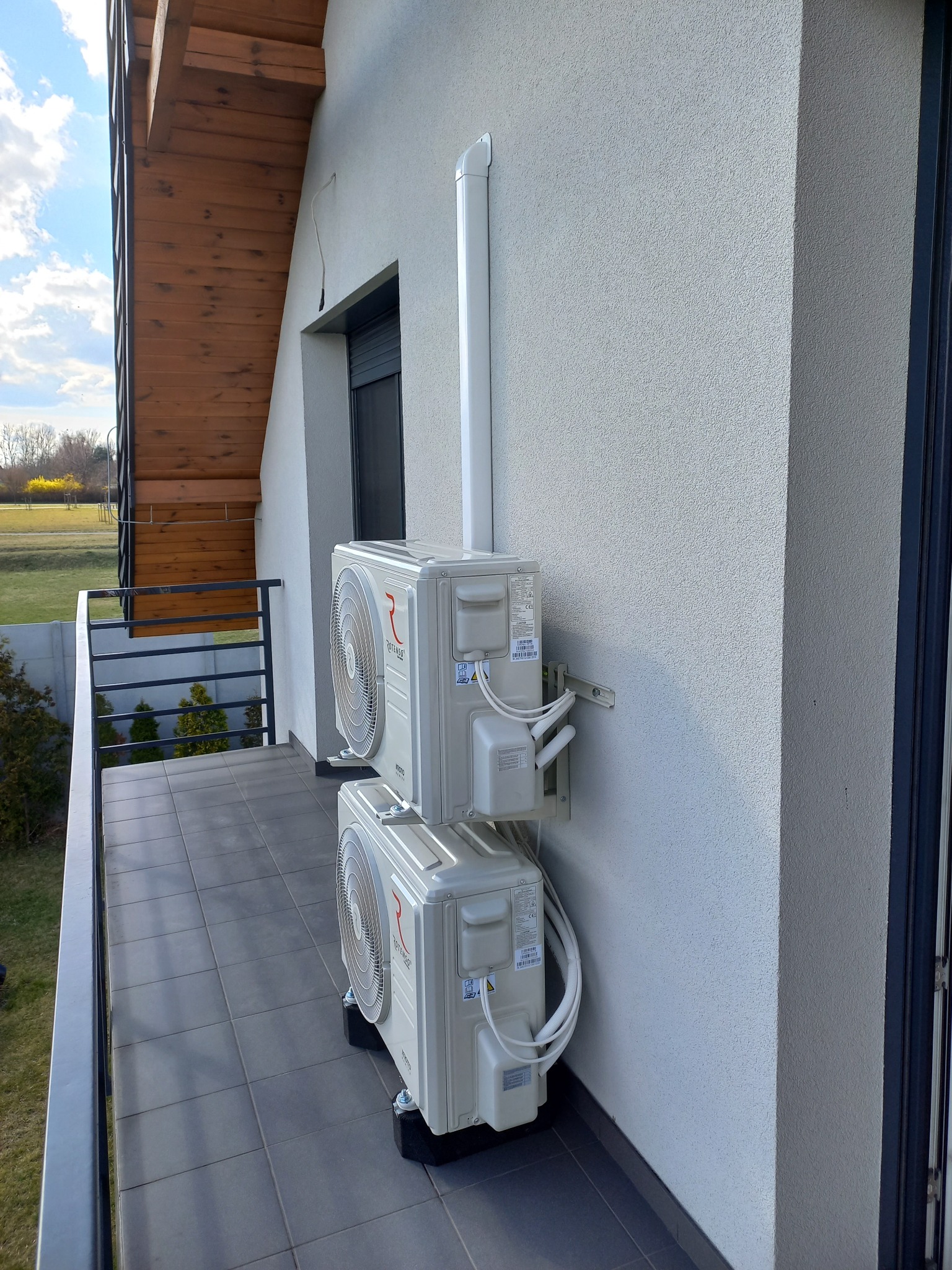 Dwa białe klimatyzatory Rotenso zamontowane jeden nad drugim na balkonie domu z widokiem na zielony trawnik i drzewa.