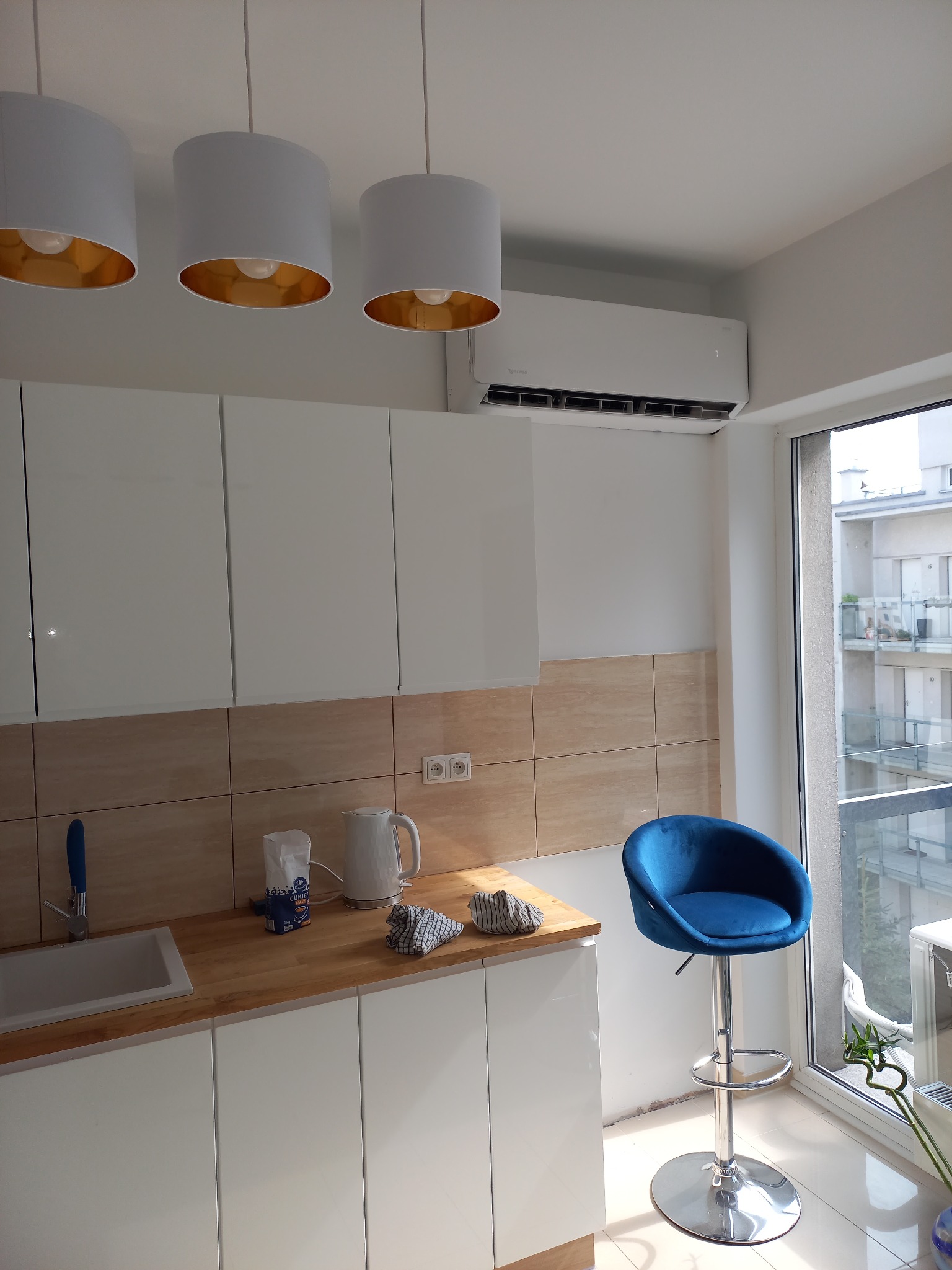 Nowoczesna, jasna kuchnia z białymi szafkami i beżowymi płytkami, oświetlona trzema lampami wiszącymi z złotym wnętrzem, z zainstalowanym klimatyzatorem ściennym nad szafkami.