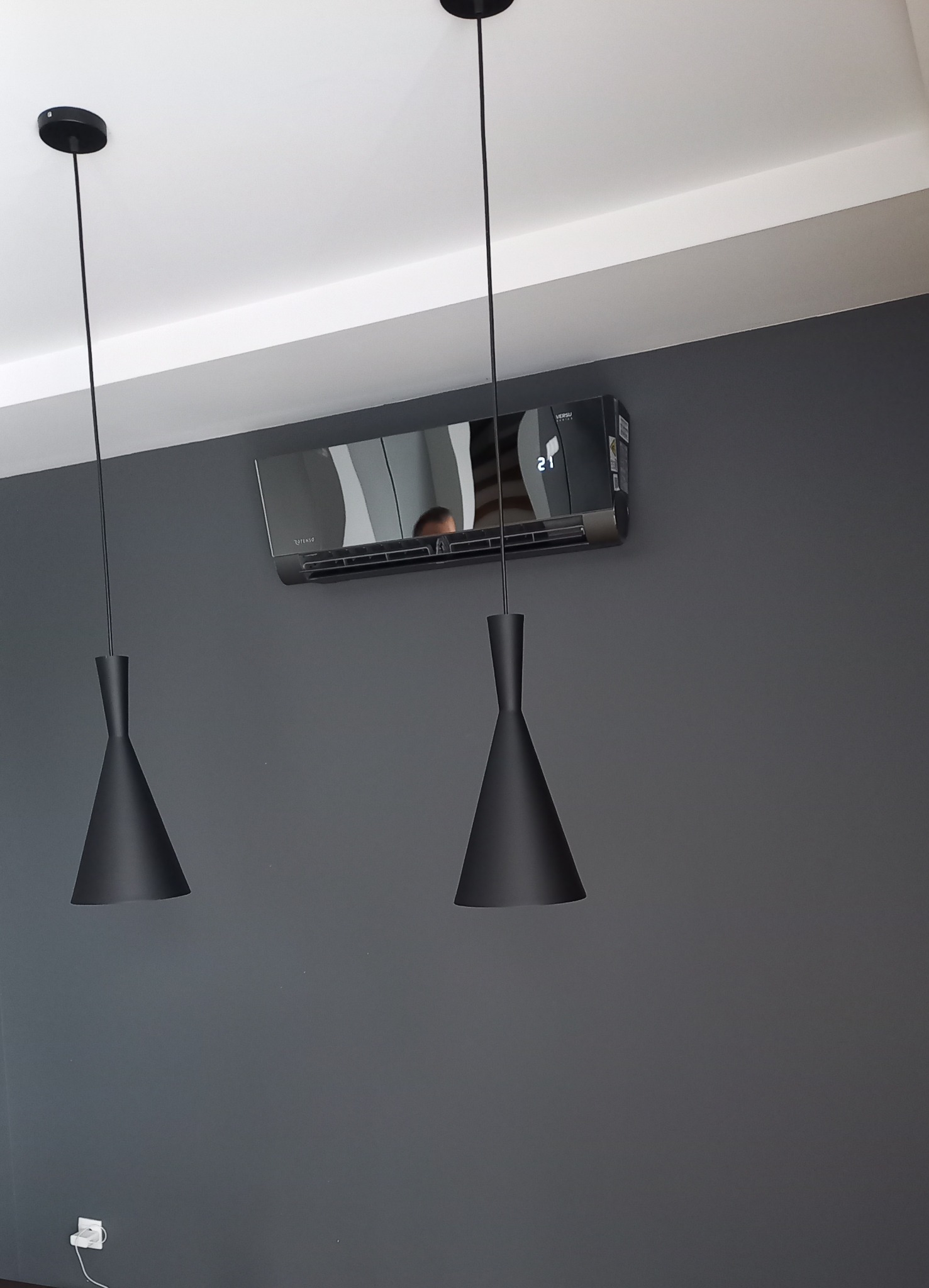 Czarny klimatyzator ścienny z wyświetlaczem temperatury, zamontowany w szarym pokoju z dwoma czarnymi lampami wiszącymi.