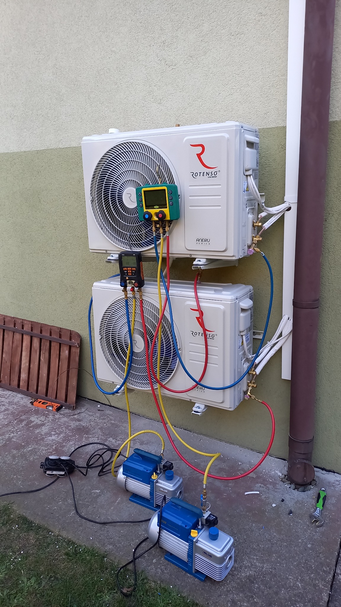 Dwa zamontowane na ścianie, jeden nad drugim, klimatyzatory marki Rotenso podłączone do urządzeń diagnostycznych i pomp próżniowych podczas serwisu. Widoczne przewody, manometry, oraz narzędzia.