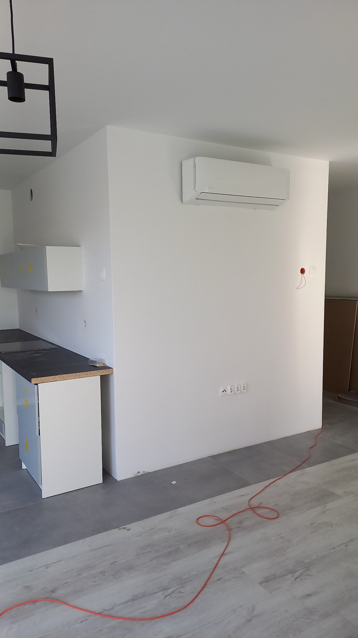 Nowoczesny klimatyzator ścienny zainstalowany w jasnym pomieszczeniu z elementami kuchennymi w trakcie montażu, widoczny kabel zasilający na podłodze.