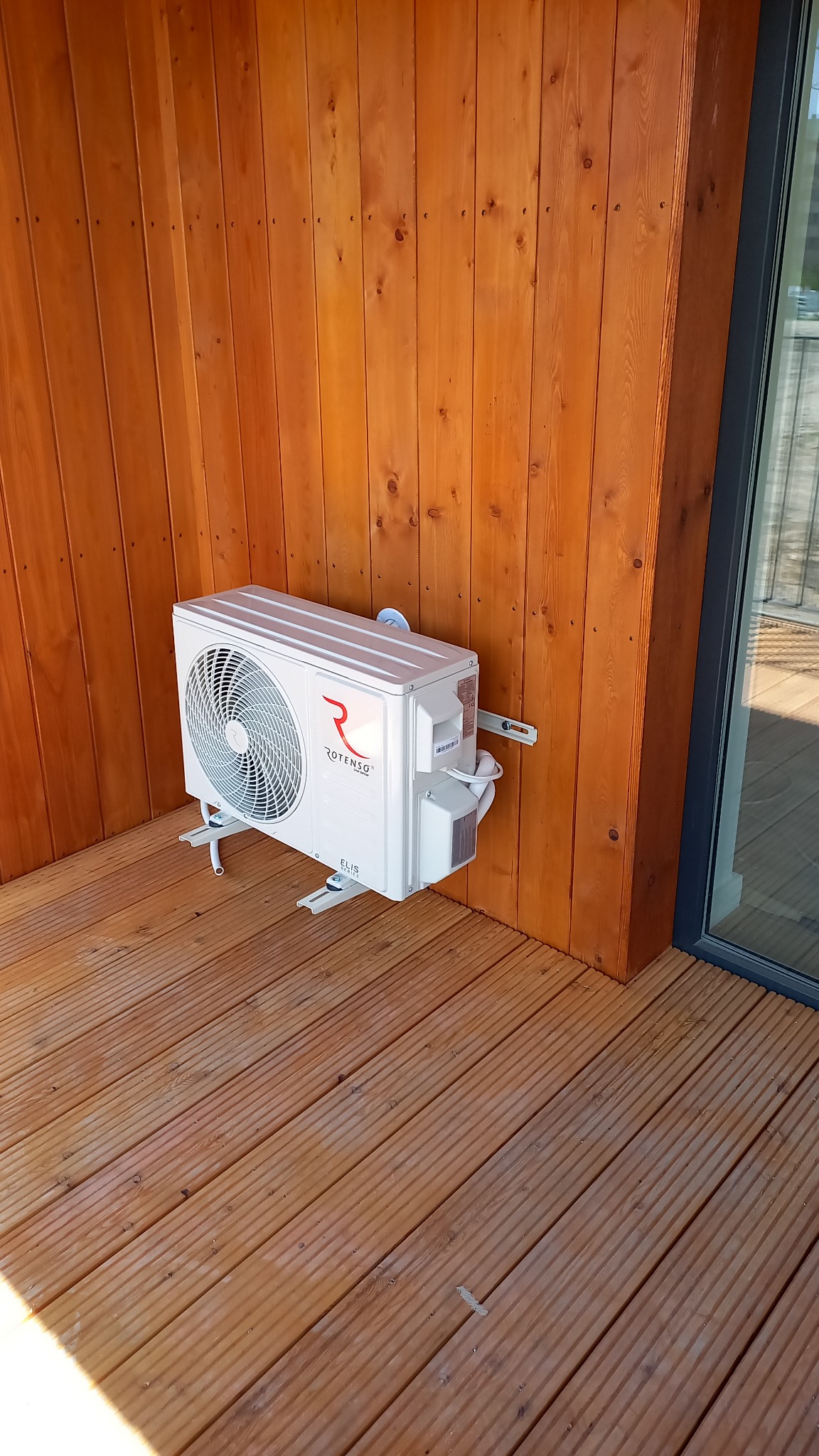 Biały klimatyzator Rotenso ELIS zamontowany na drewnianej ścianie i podłodze tarasu, widoczny z boku, z elementami montażowymi.