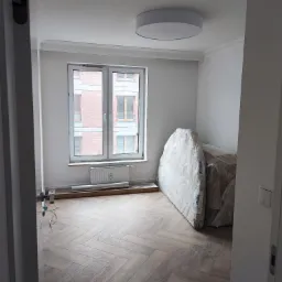 Pokój po remoncie z nową podłogą ułożoną w jodełkę, okno z widokiem na budynek z cegły, meble zabezpieczone folią, białe ściany z dekoracyjną listwą przysufitową.