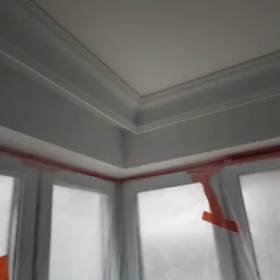 Przygotowanie do malowania: okna zabezpieczone folią i taśmą malarską, widoczny fragment sufitu z ozdobnym gzymsem.