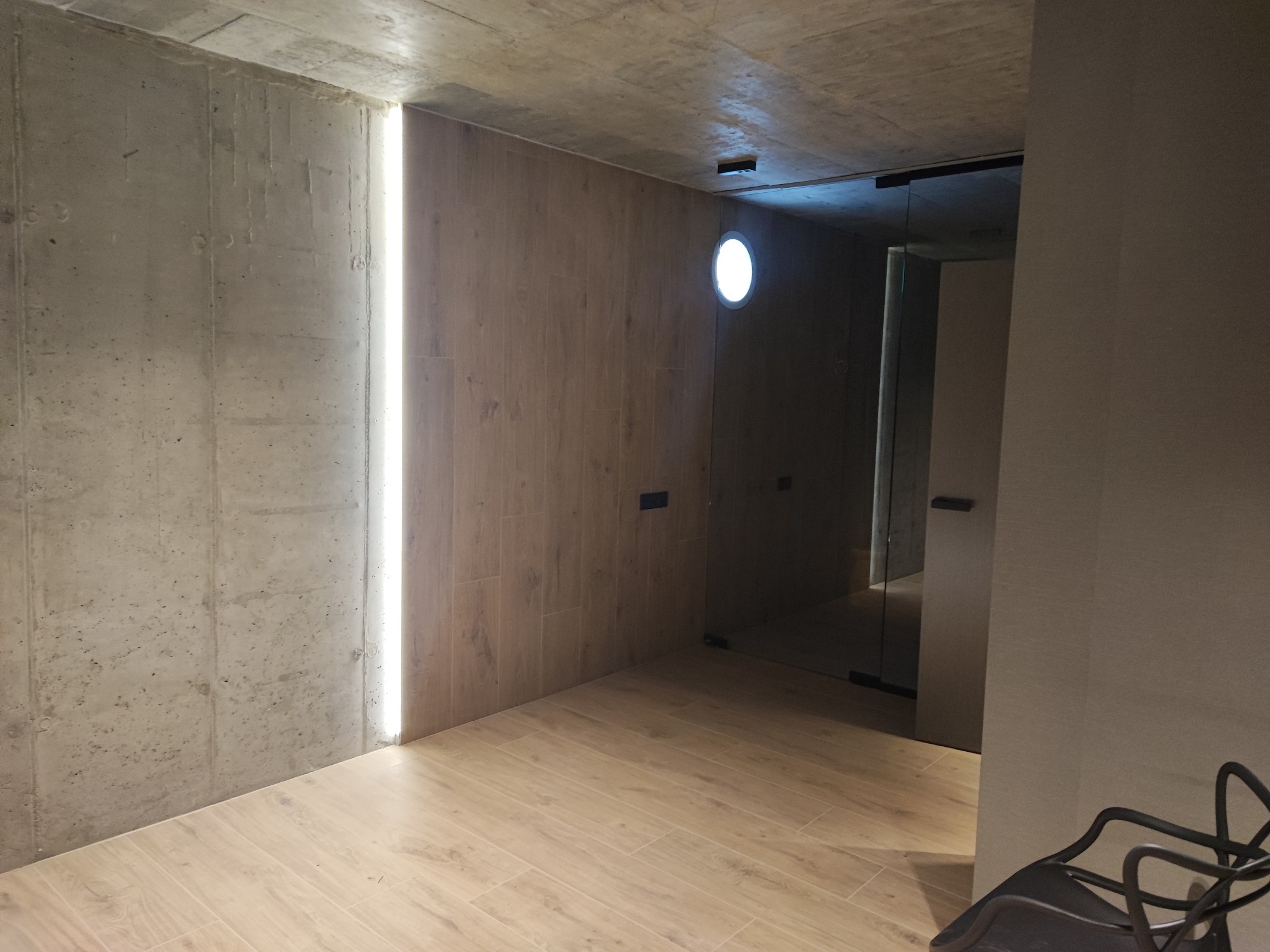 Surowe wnętrze z betonową ścianą, drewnopodobnymi panelami ściennymi i podłogą, wąską pionową lampą w ścianie, okrągłym oknem i szklanymi drzwiami do kolejnego pomieszczenia.