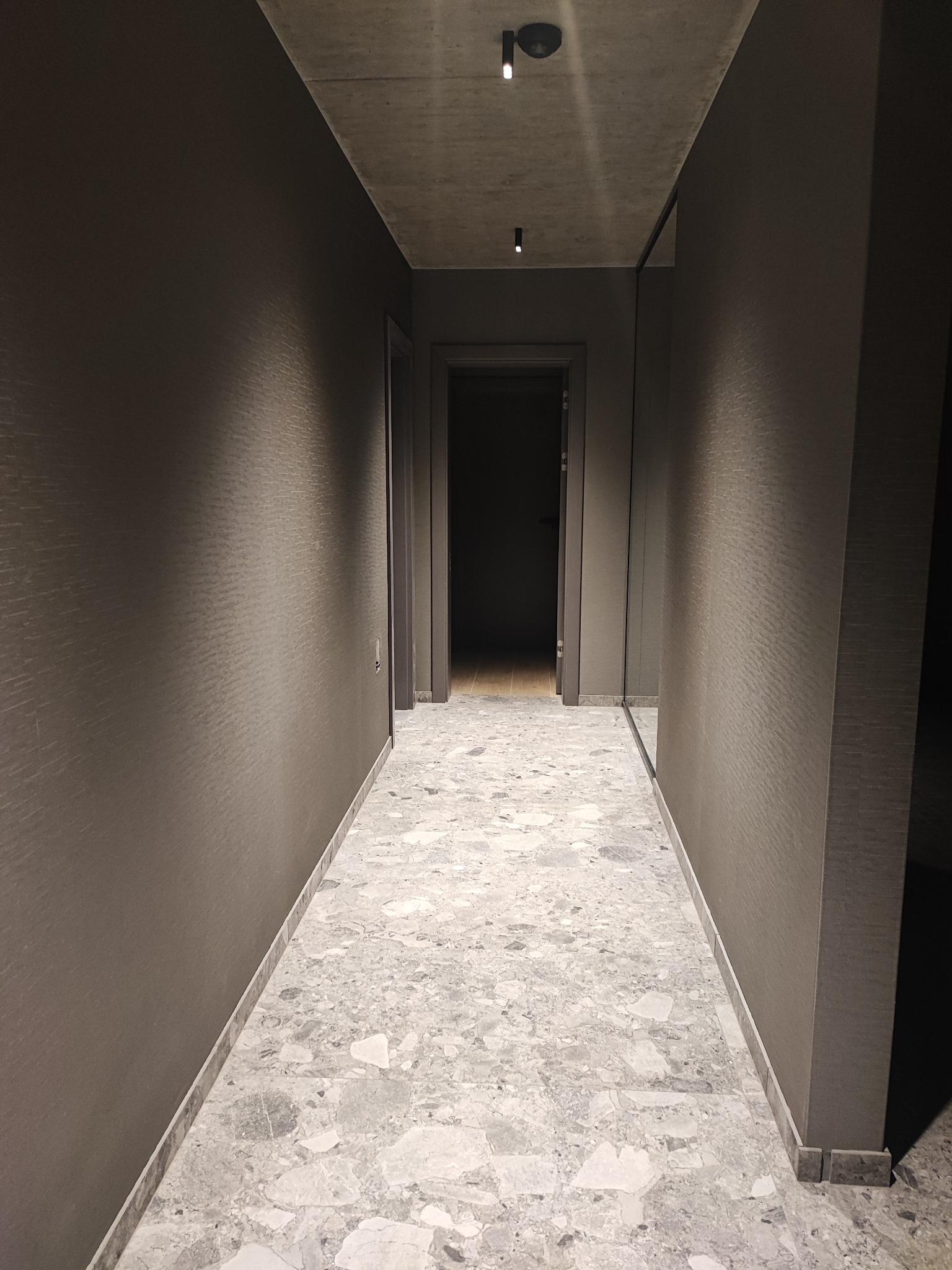 Korytarz wykończony w szarościach, z podłogą z płytek imitujących lastryko i ścianami o subtelnej teksturze, oświetlony punktowymi lampami sufitowymi.