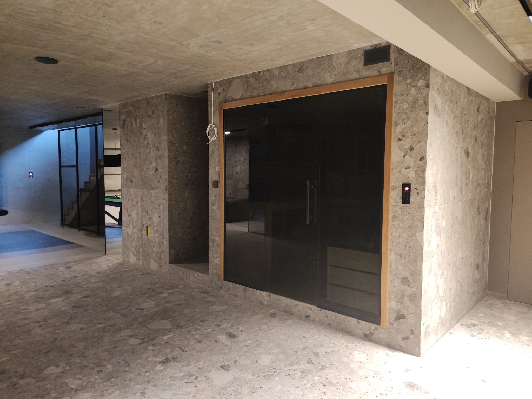 Wnętrze z wykończoną strefą sauny, ściany i podłoga wyłożone płytami w odcieniach szarości, drewniane obramowanie szklanych drzwi do sauny.