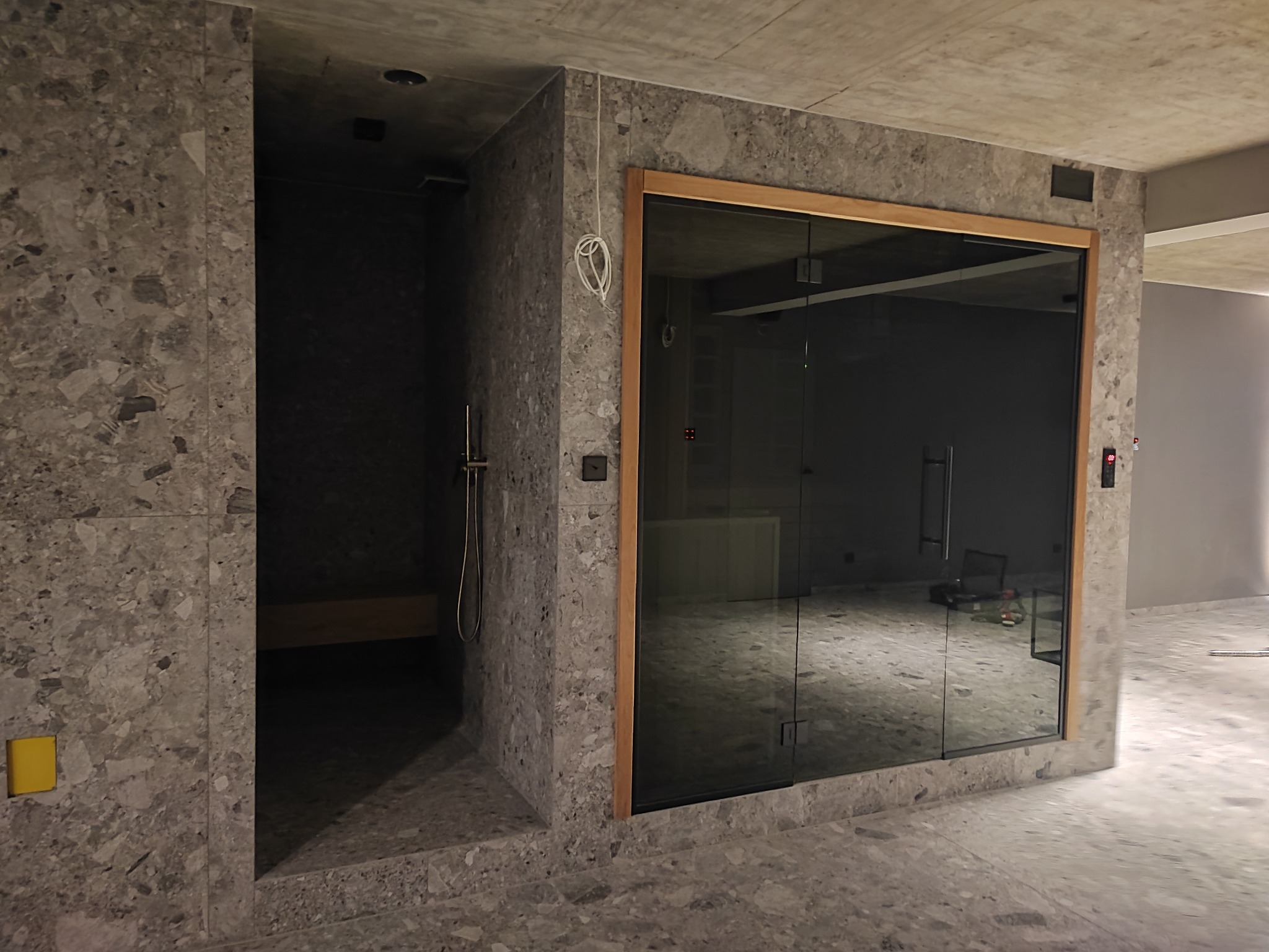 Nowoczesna łazienka w trakcie wykańczania, widoczna kabina prysznicowa z szklanymi drzwiami w drewnianej ramie oraz wnęka z półką i prysznicem, ściany i podłoga wyłożone płytkami imitującymi...