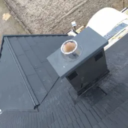 Czarny dach pokryty gontem bitumicznym z kominem wykończonym blachą, widok z góry na tle zaoranego pola.