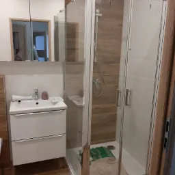 Nowoczesna łazienka z kabiną prysznicową typu walk-in, umywalką z szafką i lustrem z oświetleniem, wykończona płytkami imitującymi drewno, z widocznymi detalami jak ręczniki wewnątrz kabiny.