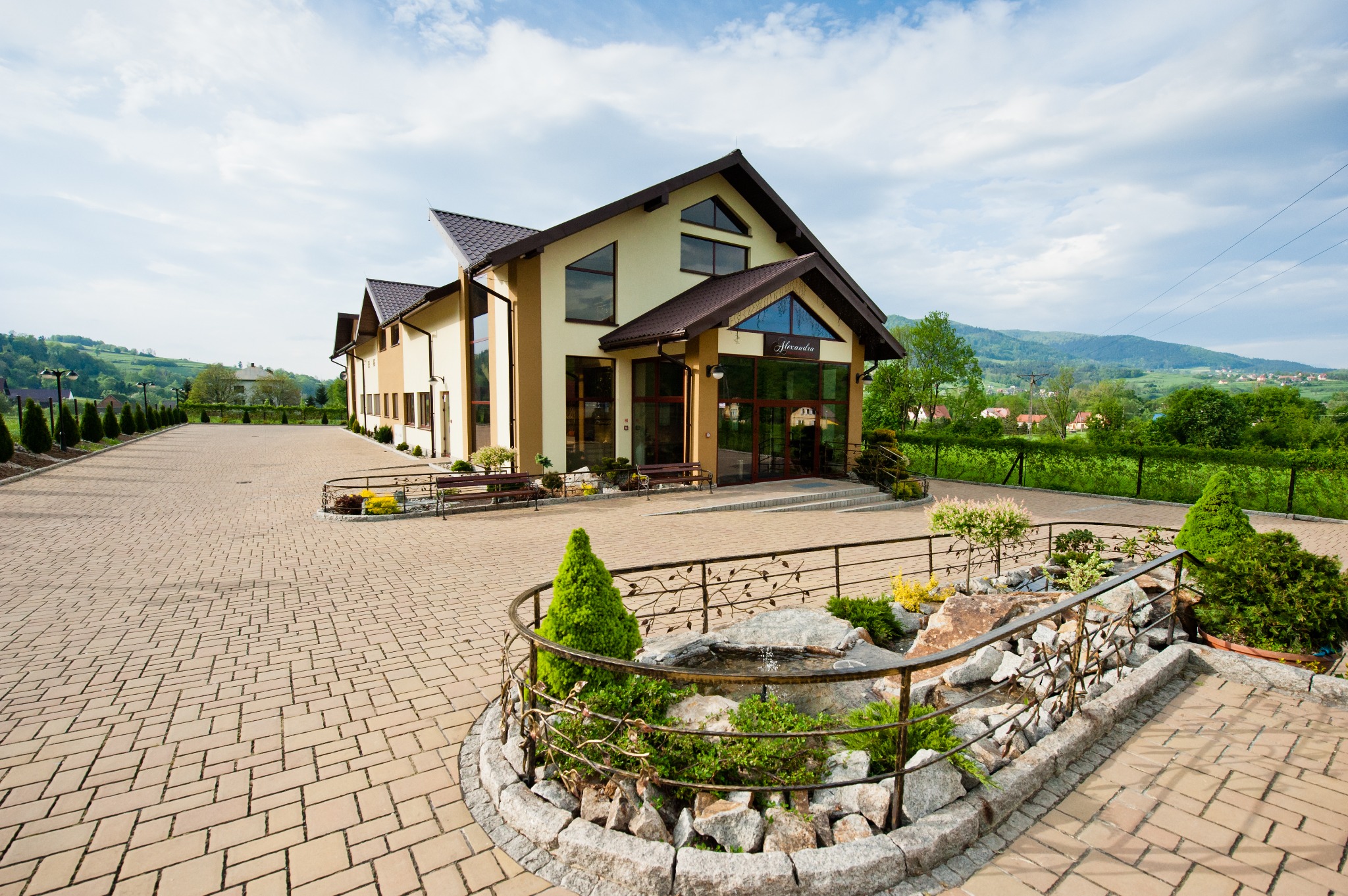 Elewacja budynku z dużymi oknami, wejście z przeszklonymi drzwiami, dekoracyjny ogródek z fontanną na pierwszym planie, w tle pagórkowaty krajobraz