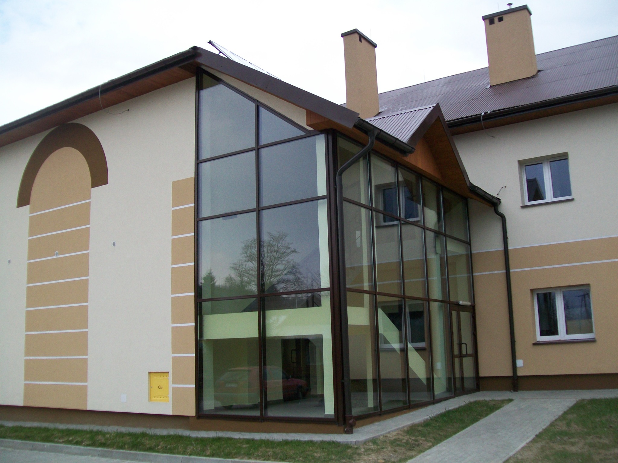 Dwukondygnacyjny budynek z dużą, przeszkloną fasadą z brązowymi ramami, odbijającą drzewa i fragment wnętrza z zaparkowanym samochodem, widok z zewnątrz na chodnik i trawnik.