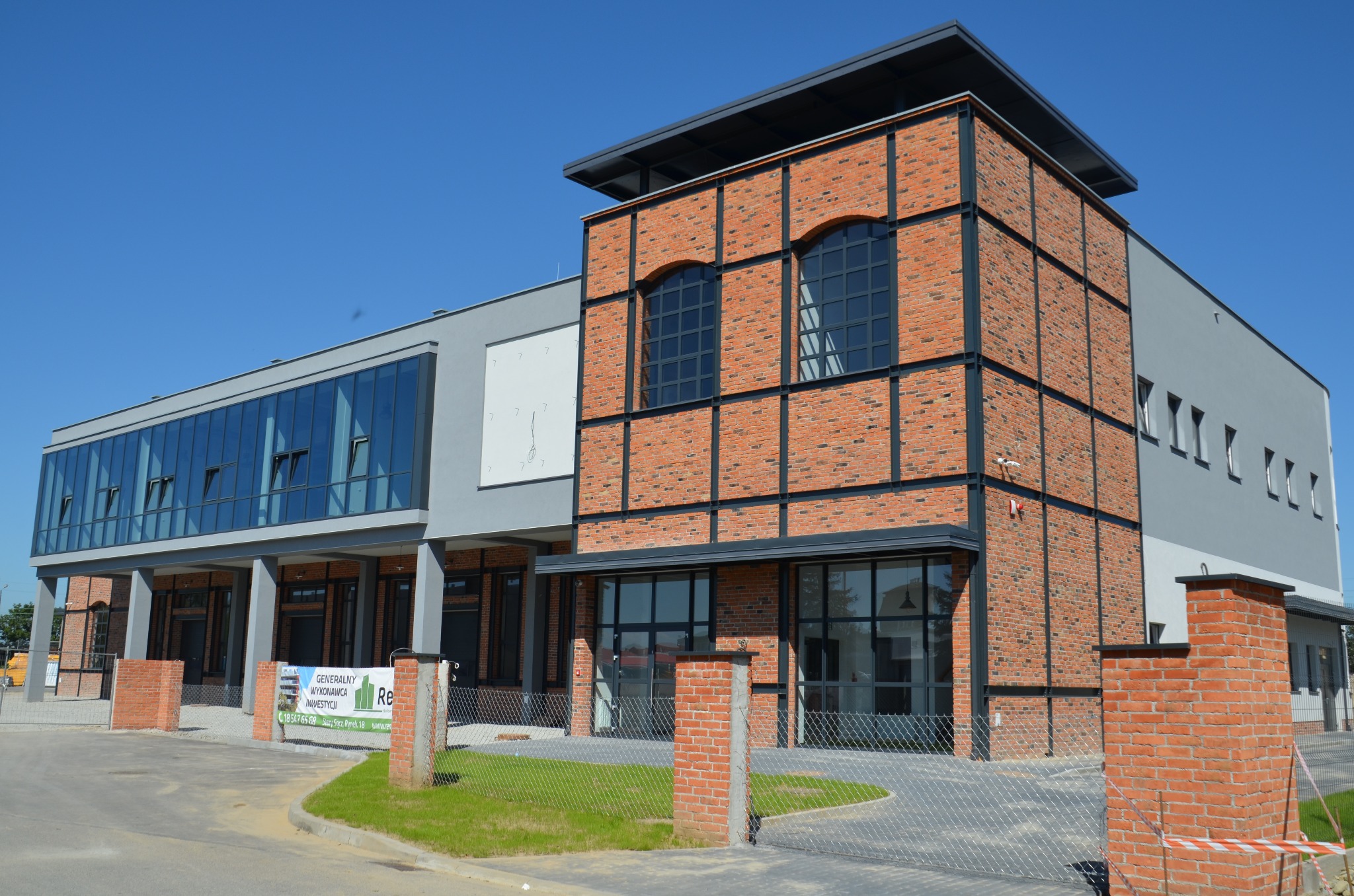 Nowoczesny budynek biurowy z ceglaną fasadą, dużymi oknami aluminiowymi z czarnymi szprosami i szarą częścią elewacji, widoczny baner reklamowy firmy budowlanej