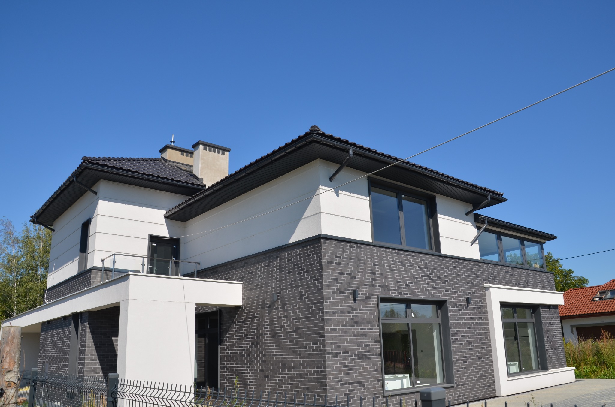 Nowoczesny dom z szarej cegły i białym tynkiem, z ciemnymi oknami i dachem, widziany z niskiego kąta pod błękitnym niebem.
