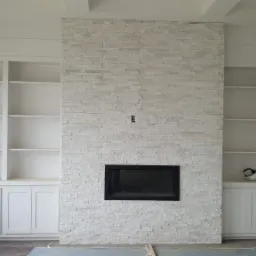 Wnętrze pokoju z kominkiem obłożonym jasnym kamieniem dekoracyjnym i wbudowanymi białymi regałami z szafkami, widoczne gniazdko elektryczne na ścianie, etap prac wykończeniowych.
