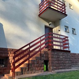 Wooden Harbor Michał Jechna - Czerwone balustrady schodów i balkonu przy jasnym budynku z ceglaną podmurówką w słoneczny dzień. Widoczny fragment trawnika.