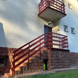 Czerwone balustrady schodów i balkonu przy jasnym budynku z ceglaną podmurówką w słoneczny dzień. Widoczny fragment trawnika.