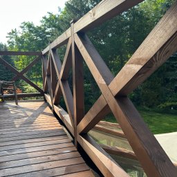 Wooden Harbor Michał Jechna - Drewniana balustrada z krzyżującymi się elementami na tarasie, w tle zieleń drzew i krzewów. Widoczne deski podłogowe i fragment mebla ogrodowego.