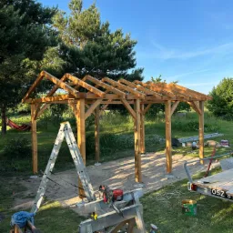 Wznoszenie drewnianej altany: konstrukcja szkieletowa z widocznymi narzędziami na trawniku, drabina oparta o filar, błękitne niebo w tle. Etap budowy.