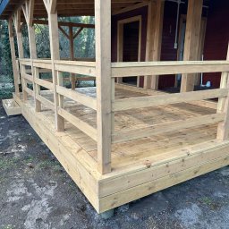 Wooden Harbor Michał Jechna - Nowo postawiona drewniana konstrukcja z zadaszeniem i barierkami, z jasnego drewna, na podmokłym terenie obok czerwonego domu. Widoczne detale łączeń i deski podłogowe.