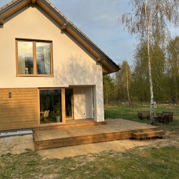 Wooden Harbor Michał Jechna - Nowoczesny dom z jasną elewacją i drewnianymi panelami, z rozległą drewnianą platformą zewnętrzną. W tle brzozy i las, oświetlone ciepłym światłem zachodzącego słońca.