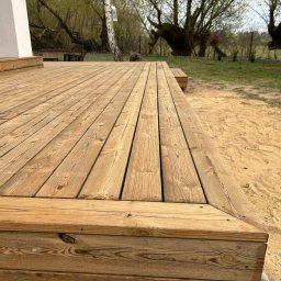 Wooden Harbor Michał Jechna - Solidny podest z jasnych, naturalnych desek, z widocznymi słojami i wkrętami, tworzący narożnik. Obok piaszczysty grunt i zieleń z brzozą z lampionem.